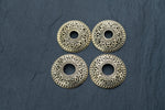 On a dark, textured background, four intricately designed circular pieces from the Kaya Movement collection are arranged in a square formation. These gold-colored items, known as Gold brest buttons piercing jewelry rings and teat body jewelry shields, resemble ornate buttons with a central hole and could be used as decorative elements or jewelry components.