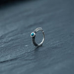 A delicate 925 silver Indian nose ring by Kaya Movement lies on a dark, textured surface. The ring features an 8mm circular turquoise stone set within a bezel, with an intricate, beaded design along the band. Perfect for septum, rook piercings, tragus piercings, or helix jewelry enthusiasts, the overall aesthetic is minimalistic yet elegant.