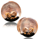 The Handmade Epoxy Double Flare Flesh Tunnels with Skull and Gothic Design by Kaya Movement feature two round plugs encapsulating small skulls in clear, swirling resin on a base of dark material adorned with gold flecks. This gauged ear stretching jewelry, available in sizes from 12mm to 16mm, exudes a haunting yet artistic appearance, making it uniquely striking.