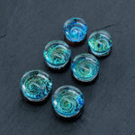 A set of six handmade Blue Cosmic Ear Plugs from Kaya Movement, featuring dome-shaped glass cabochons with mesmerizing blue and green swirls resembling galaxies. These unique celestial body jewelry pieces for stretching, available as spiral gauges and double flare tunnels in 12mm-16mm sizes, are arranged on a dark, textured surface to showcase their enchanting beauty.