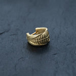 A single 18k gold-plated septum ring with a textured, biomechanical alien design from Kaya Movement sits on a dark, slate surface. The Futuristic Xenomorph Septum Ring has an open back, allowing it to be easily adjusted and worn without the need for a piercing. The background contrasts elegantly with the shiny gold metal, highlighting the intricate details meant for both men and women.