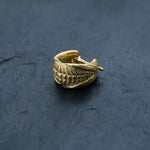A close-up of the Kayа Movement's Futuristic Xenomorph 18k Gold-Plated Septum Ring for Men and Women, 16G, on a dark, textured surface. The septum ring features an intricate design resembling a series of overlapping leaves or scales, giving it a detailed and elegant appearance akin to a biomechanical horseshoe design.