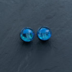 The Blue Glass Jellyfish Ear Plugs by Kaya Movement feature an intricate Blue Jellyfish design encased inside small, round plugs. The jellyfish appear to float in a deep, translucent blue resin, creating a captivating 3D effect against a dark grey textured background. These handmade double flare flesh tunnels are available in sizes ranging from 12mm to 16mm and make stunning ocean-themed body jewelry that captivates and enchants.