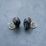 The Handmade Black Skull Dangle Earrings by Kaya Movement feature skull-shaped pendants that hang from gold hooks, set against a textured gray surface. The intricate details of this gothic tribal jewelry are highlighted by the contrasting dark hue of the black skulls against the gold.