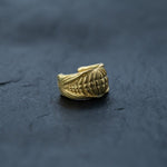 A gold-colored, intricately designed ring with a seashell motif sits on a dark, textured surface. The Kaya Movement Futuristic Xenomorph 18k Gold-Plated Septum Ring for Men and Women features a detailed pattern that mimics the natural lines and curves of a seashell, creating an elegant and unique piece of jewelry.