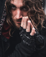 A person with long, wavy hair and a nose ring gazes intently at the camera. They are wearing Kaya Movement's Custom Dragon Scale Leather Gloves, which are fingerless motorcycle and driving gloves perfect for post-apocalyptic goth stagewear. Along with a hooded jacket adorned with metallic studs, their hands partially cover their face, adding a mysterious and intense expression that hints at gothic fashion influences.