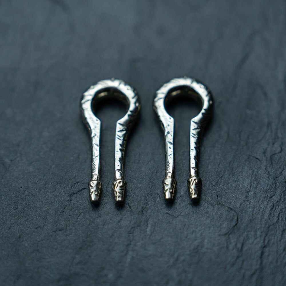 Two traditional jaw harps with textured metal designs are placed side by side on a dark, textured surface. Each harp has a looped structure with a straight, narrow tongue extending from the base, showcasing intricate craftsmanship that is reminiscent of the detailed work found in Kaya Movement's Silver White Brass Ear Weights for stretched lobes body mod jewelry.