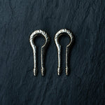 Two intricately designed ear weights from Kaya Movement, crafted in silver white brass and shaped like keyhole hangers for stretched lobes, resting side by side on a dark, textured surface.