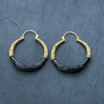 On a textured dark surface lies a pair of Handmade Boho and Tribal Horn Earrings from Kaya Movement. Each earring features an intricately patterned gold upper part and a black lower part carved from ethically sourced natural buffalo horn, creating a striking contrast that adds an elegant touch to these cool statement earrings.