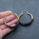 A hand holding a pair of striking Handmade Boho and Tribal Horn Earrings from Kaya Movement. These gold-plated earrings, adorned with intricate swirling designs, feature black, crescent-shaped embellishments reminiscent of natural buffalo horn. At the base, carved patterns add an elegant touch against the dark, textured background.