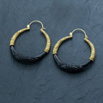A pair of stunning Handmade Boho and Tribal Horn Earrings from Kaya Movement are displayed on a dark slate background. Each earring features a half-gold, half-black design, with intricate floral patterns adorning the gold upper half and a smooth black lower half. These unique hoops, part of our ethically sourced jewelry collection, are connected by small gold clasps.