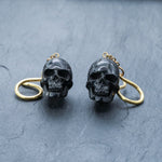 The Handmade Black Skull Dangle Earrings by Kaya Movement, featuring intricate designs and gothic tribal style, are displayed on a textured gray surface. Each earring hangs from a looped gold-colored hook, adding a striking statement to the detailed skull jewelry.