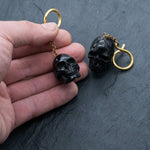 A hand holding a keychain featuring a small, black skull with another identical keychain lying nearby on a dark, textured surface. The keychains, resembling the Handmade Black Skull Dangle Earrings - Gothic Tribal Jewelry for Statement Style from Kaya Movement, have gold-colored loops and chains attached to the skulls.