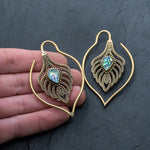 Close-up image of a hand holding ornate Kaya Movement Unisex Handmade Brass Leaf Ear Weights with Abalone Shell, designed in the shape of peacock feathers and adorned with intricate detailing. These Boho Hippie Tribal Earrings feature an iridescent gemstone resembling abalone shell and the hand provides a sense of scale for this edgy urban fashion jewelry.