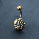 Introducing the Gold Lotus Flower Belly Ring from Kaya Movement. This stunning piece features a teardrop-shaped zircon gemstone elegantly surrounded by lotus petals. The 9mm belly bar is complemented with a small, spherical bead at the top, and is displayed beautifully against a dark textured background. Perfect for adding a touch of sophistication to your navel jewelry collection.