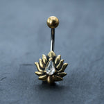 A gold Lotus Flower Belly Ring by Kaya Movement, featuring a zircon gemstone teardrop piercing, is centered on a slate-gray surface. This elegant navel jewelry includes a 9mm belly bar with a gold sphere at the top and a clear zircon gemstone, teardrop-shaped, gracefully hanging from the center of the lotus flower design.