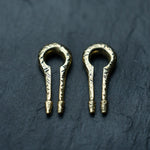 A pair of ornate, metallic objects resembling miniature keys with rounded tops and two prongs at the bottom, resting on a dark, textured surface. These Kaya Movement Gold Ear Weights in Keyhole & Hanger Style feature intricate engravings and could be mistaken for exquisite body mod jewelry designed for stretched lobes.