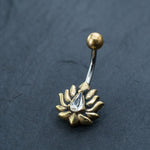 The Kaya Movement Gold Lotus Flower Belly Ring is a stunning piece of navel jewelry featuring a delicate floral design and centered with a teardrop-shaped zircon gemstone. This exquisite 9mm belly bar is beautifully displayed against a dark, textured background.
