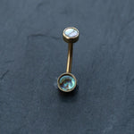 A Kaya Movement gold-toned curved barbell earring, featuring two round ends with shimmering abalone shell inlays, resembles an elegant piece from their boho-chic collection of 18k navel piercing and gold navel rings. This discreet, hypoallergenic belly jewelry is artfully displayed on a dark, textured surface.
