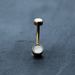 An 18k gold navel ring from Kaya Movement, featuring boho-chic, hypoallergenic MOP shell stones at each end, stands upright on a smooth, dark blue-gray surface. The sleek and polished appearance of this discreet body jewelry is highlighted against the contrasting background.