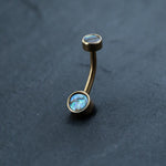 The 18k Navel Piercing & Gold Navel Ring by Kaya Movement, designed as exquisite belly jewelry in a boho-chic style, features two iridescent abalone shell rings resting on a dark, textured surface.