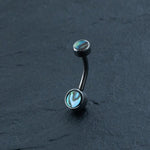 A barbell-style curved belly button ring from Kaya Movement lies on a dark, textured surface. This Boho-Chic piece from their Belly Button Jewelry collection features vibrant green and blue iridescent abalone and shell gemstone ends. Made of hypoallergenic Surgical Steel 316L, the silver metal contrasts beautifully with the gemstones, making this discreet navel piercing an eye-catching addition to your body jewelry lineup.