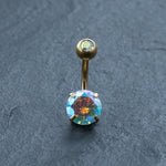 The Kaya Movement 18K Gold Navel Ring features a hypoallergenic crystal belly barbell with a small iridescent gem on the curved barbell and a larger multicolored gem at the base. Displayed on a dark textured surface, it showcases this stunning piece of allergy-friendly body jewelry.