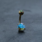The Kaya Movement 18K Gold Navel Ring, also known as the Crystal Belly Barbell, is displayed against a dark textured background. This hypoallergenic and allergy-friendly body jewelry features an opalescent gem at each end. The larger bottom gem is multi-faceted and reflects a stunning array of blue, green, and yellow hues, offering both style and comfort for discreet piercing enthusiasts.