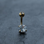 The Kaya Movement 18K Gold Navel Ring is a hypoallergenic belly barbell, adorned with a small crystal on top and a larger cubic zirconia accent on the bottom, gracefully displayed against a textured dark gray background.