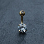 The 18K Gold Navel Ring by Kaya Movement features a small diamond at the top and a larger, multifaceted diamond at the bottom, accented with cubic zirconia. This hypoallergenic and discreet belly barbell is elegantly displayed on a dark, textured surface.