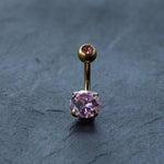 The Kaya Movement 18K Gold Navel Ring, known for its elegance and comfort, features two pink gemstones—a larger one at the bottom and a smaller one embedded in the top ball—set against a dark textured surface. This hypoallergenic belly barbell is perfect for those seeking discreet, allergy-friendly body jewelry.
