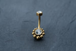The Kaya Movement 18K Gold Navel Ring, named Crystal & CZ Belly Barbell, features clear cubic zirconia accents on each end. The larger gemstone is encircled by small gold beads in a circular pattern, adding an elegant touch. This hypoallergenic and discreet belly barbell is beautifully displayed on a dark, textured surface.