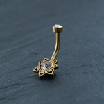 The 18K Gold Navel Ring from Kaya Movement showcases a clear cubic zirconia gemstone at the top and a decorative charm at the bottom. The charm features a larger central gemstone encircled by intricate patterns with small heart shapes. This Crystal & CZ Belly Mandala Barbell, known for its hypoallergenic properties and discreet design, rests elegantly on a dark slate surface.