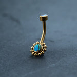 A stunning 18k navel piercing from Kaya Movement, this gold belly button ring features a curved bar with a circular abalone shell surrounded by delicate gold beads. An additional small, flat stud graces the top. The background is a smooth, dark surface showcasing this exquisite piece of boho-chic style and hypoallergenic body jewelry.