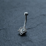 The Hypoallergenic Crystal Zircon Belly Ring by Kaya Movement is a discreet CZ navel piercing crafted from allergy-friendly Surgical Steel 316L Stainless in a silver tone. It features a round, clear gemstone at one end and is adorned with three small black heart-shaped decorations. This elegant piece of belly button jewelry lies on a dark, textured surface.