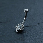 A silver Hypoallergenic Crystal Zircon Belly Ring by Kaya Movement with a curved barbell lies on a dark surface. It features a central round clear gemstone, surrounded by an ornate design with small heart shapes at its edges. The top end of the Surgical Steel 316L Stainless barbell is adorned with another clear gemstone.