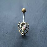 An Abalone Shell Belly Button Ring by Kaya Movement, featuring a 9mm surgical steel bananabell with an intricate boho design and an iridescent stone in the center, surrounded by a geometric metallic pattern, is placed on a dark, textured surface.