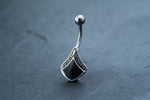 Close-up of a Black Pearl Belly Ring by Kaya Movement against a dark, textured background. The silver navel tribal bar showcases an elegant, curved design featuring a polished, black triangular stone encased in a silver frame with intricate patterns, evoking the charm of an ethnic-inspired piece.
