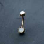 A curved, gold-tone barbell piercing with round white MOP shell stones set in both ends, displayed on a dark, textured surface. The minimalist design gives this Kaya Movement 18k Navel Piercing & Gold Navel Ring a modern, elegant look.