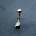 An 18k gold navel ring from Kaya Movement, adorned with round white MOP shell stones on each end, is placed vertically against a dark gray textured background.