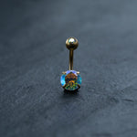 A close-up of the Kaya Movement 18K Gold Navel Ring, Crystal Belly Barbell highlights its iridescent round gemstone. The gemstone beautifully reflects a spectrum of colors, with predominant hues of blue, green, and orange when light strikes it. This hypoallergenic and discreet piece of body jewelry stands out against a dark, textured background.