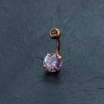 The Kaya Movement 18K Gold Navel Ring, featuring a hypoallergenic pink crystal belly barbell with a curved design, showcases two pink gemstones set in gold-colored metal against a dark textured background. The larger gemstone is positioned at the bottom while the smaller one sits at the top, making this discreet and allergy-friendly body jewelry both elegant and safe for sensitive skin.