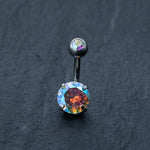 Displayed on a dark, textured background is the Kaya Movement’s Discreet Allergy-Friendly Crystal Belly Barbell - a shimmering surgical silver navel ring featuring a large, multicolored iridescent zircon. This sparkling piece of body jewelry showcases an array of colors including red, blue, and green for an eye-catching effect and is perfect for belly button piercings.