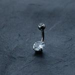 A close-up of the Hypoallergenic Crystal Zircon Belly Ring from Kaya Movement showcases the Allergy-Friendly Silver Steel Navel Piercing. This discreet CZ belly button jewelry, made from surgical steel 316L, features a silver curved barbell adorned with a small round crystal on one end and a larger oval crystal on the other, set against a dark textured background.