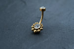 Here's a rephrased sentence using the given product data:
The Kaya Movement 18K Gold Navel Ring, adorned with two clear gemstones, offers stunning elegance. The bottom gemstone is encircled by a ring of gold beads for added sophistication, while the smaller top gemstone is set in a simple gold setting. With cubic zirconia accents and hypoallergenic properties, this discreet belly barbell rests beautifully on a dark, textured surface.