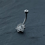 A close-up of the Kaya Movement Hypoallergenic Crystal Zircon Belly Ring shows a beautiful flower design with a clear crystal at its center. Each heart-shaped petal is also adorned with small crystals, set against a dark, textured background. This discreet CZ navel piercing is allergy-friendly and made from silver surgical steel.