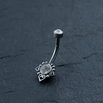 The Kaya Movement Hypoallergenic Crystal Zircon Belly Ring features a curved metal design made from Surgical Steel 316L Stainless, with a clear gemstone at one end. The other end showcases a decorative arrangement, including another clear gemstone surrounded by small metal beads and heart shapes. This allergy-friendly silver steel belly button jewelry is set against a dark, textured surface to enhance its intricate details.