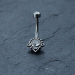 The Kaya Movement Hypoallergenic Crystal Zircon Belly Ring, featuring a smaller clear gemstone at the top and a larger one at the bottom, both surrounded by heart-shaped decorations, is showcased on a dark textured surface. Crafted from Surgical Steel 316L Stainless for durability and style, this discreet CZ navel piercing offers an allergy-friendly option for silver steel belly button jewelry enthusiasts.