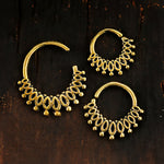 Displayed on a dark wooden surface is a unique set from Kaya Movement: The Gold-Plated Brass Septum in Maya Tribal Design. This exquisite piece, suitable for nose, helix 1.2mm, tragus, lobe, and conch piercings, embodies an intricate bohemian jewelry style with delicate leaf motifs and bead-like elements edging the design. The largest septum hoop is positioned on the left, accompanied by two smaller hoops to its right.