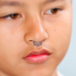 A close-up showcases a person with tan skin wearing Kaya Movement's elegant Fake Septum "Solara" 925 Silver Mandala. The no-pierce nose jewelry features a detailed and intricate mandala design crafted from pure silver. The person's facial expression is neutral, while the background remains blurred.